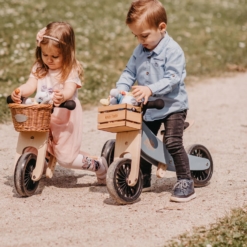 Two children, a boy and a girl ride Kinderfeets Tiny Tots in a sunny field with a gravel road. The boy's Tiny Tot has a wooden crate accessory, while the girl's Tiny Tot has a woven basket on the front.